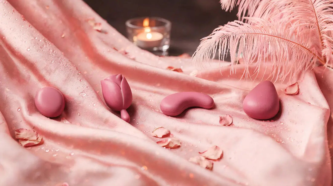 Four rose-pink waterproof intimate-wellness devices on pink silk with ostrich feathers and candle bokeh