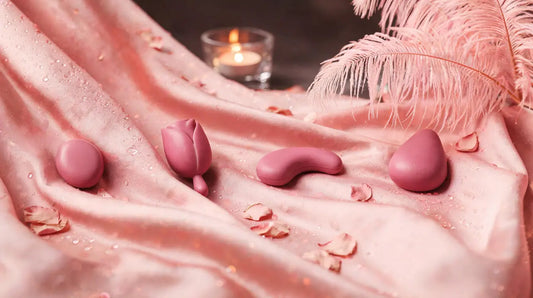 Four rose-pink waterproof intimate-wellness devices on pink silk with ostrich feathers and candle bokeh