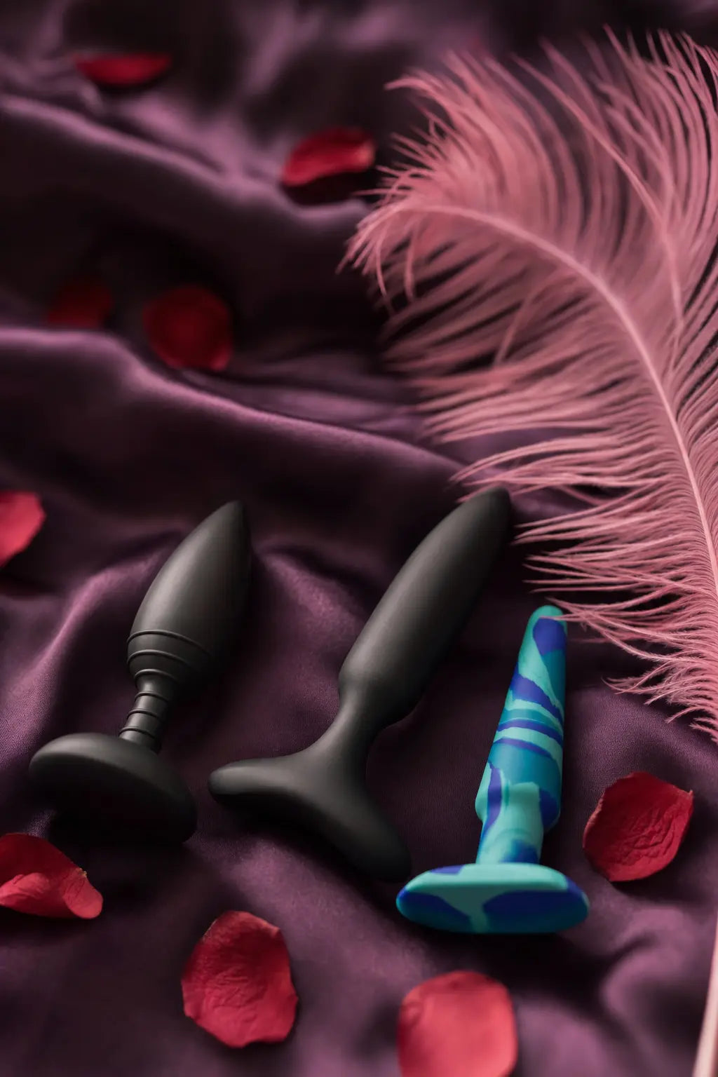 Editorial flat-lay of beginner-friendly butt plugs on aubergine silk with pink feather