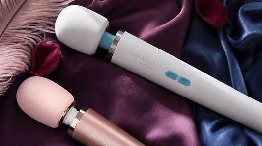 Editorial flat-lay featuring Le Wand Classique Rechargeable and Le Wand Petite wand massagers, purple silk, pink feather