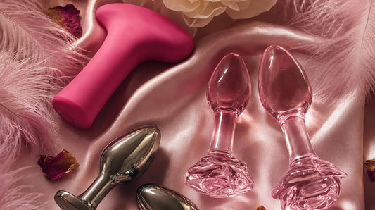 Flat lay of silicone, borosilicate glass and stainless steel pleasure products on blush-pink satin with pink feathers, rose petals and a white peony, lit by warm candlelight