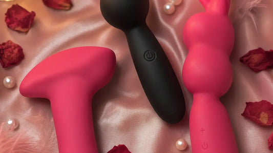 Three soft silicone vibrators — a bullet, a mini wand, and a bunny wand — resting on blush-pink satin with rose petals, pearl beads, and a glowing candle in the background