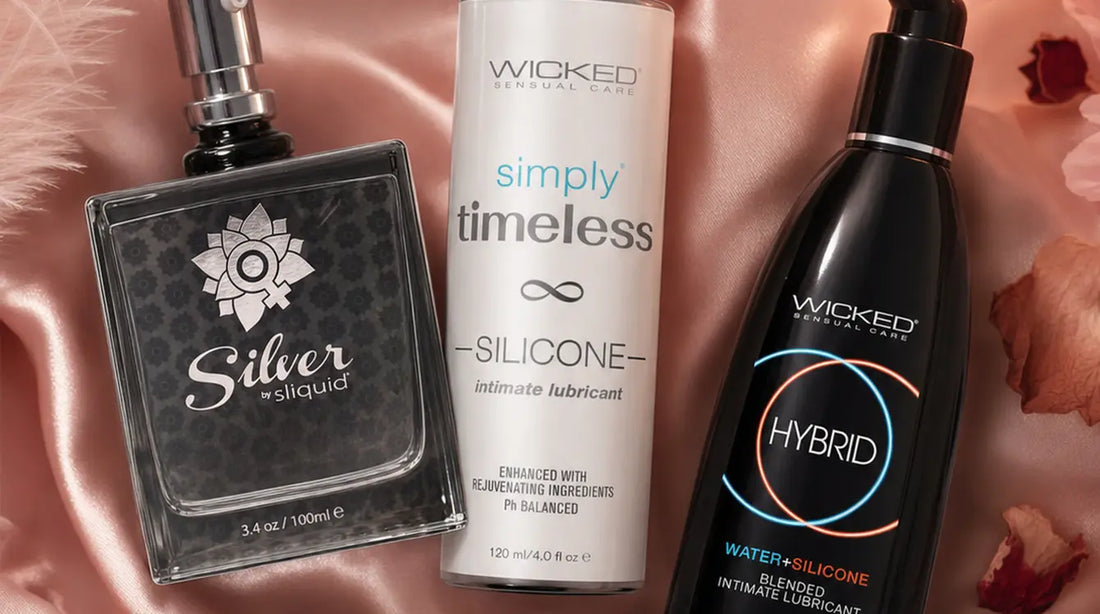 Three lubricant bottles — Sliquid Silver, Wicked Sensual Care Timeless Silicone, and Wicked Hybrid — arranged on dusty rose silk satin with pink feathers, rose petals, and a glowing candle