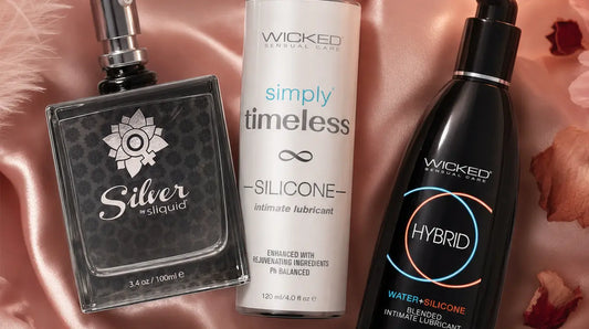 Three lubricant bottles — Sliquid Silver, Wicked Sensual Care Timeless Silicone, and Wicked Hybrid — arranged on dusty rose silk satin with pink feathers, rose petals, and a glowing candle