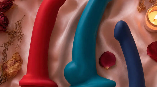 Three platinum silicone suction-cup dildos in crimson, teal, and indigo arranged on blush pink satin fabric with rose petals, dried flowers, and soft candlelight