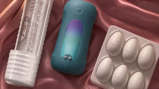 Three male masturbators — a white cylindrical sleeve, a teal handsfree stroker, and a pack of egg-style sleeves — arranged on rose-pink satin with pink feathers and dried flower petals, lit by soft candlelight