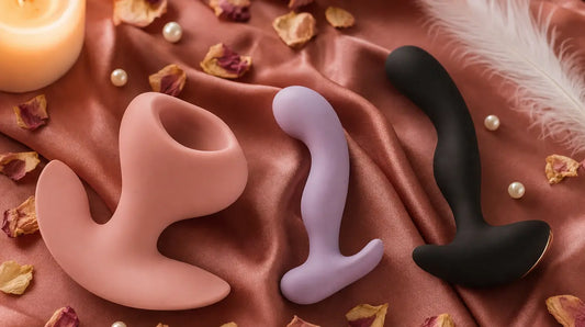 Three silicone intimate wellness products arranged on rose-gold satin fabric with dried rose petals, pearl beads, a white feather, and soft candlelight