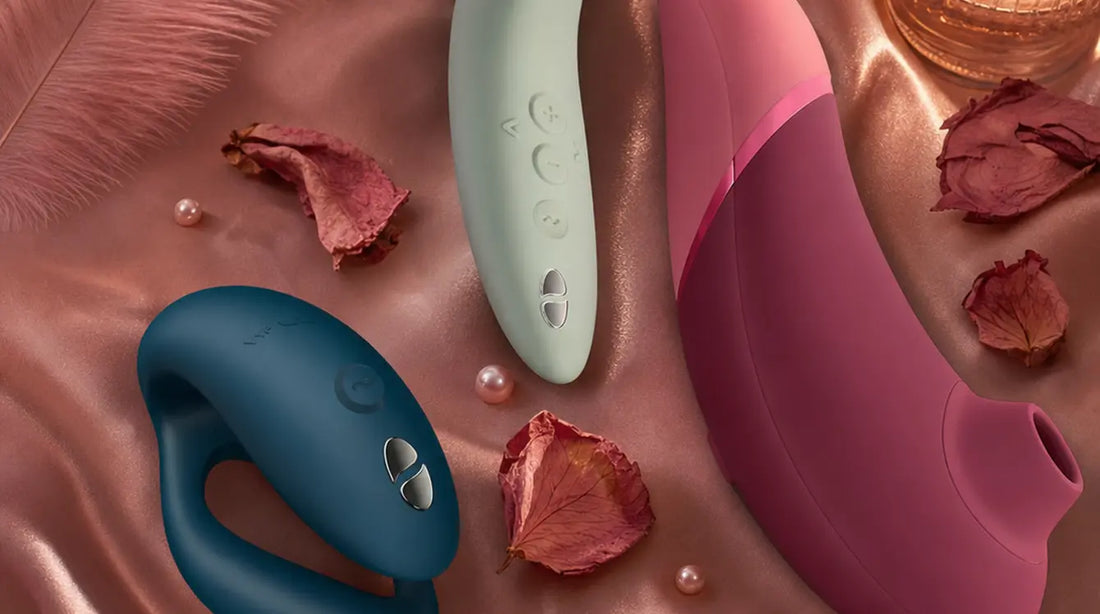 Three premium pleasure devices — a teal couples ring, a mint green stimulator, and a deep pink pleasure air toy — arranged on blush satin fabric with scattered rose petals, pearls, and pink feathers in warm candlelight