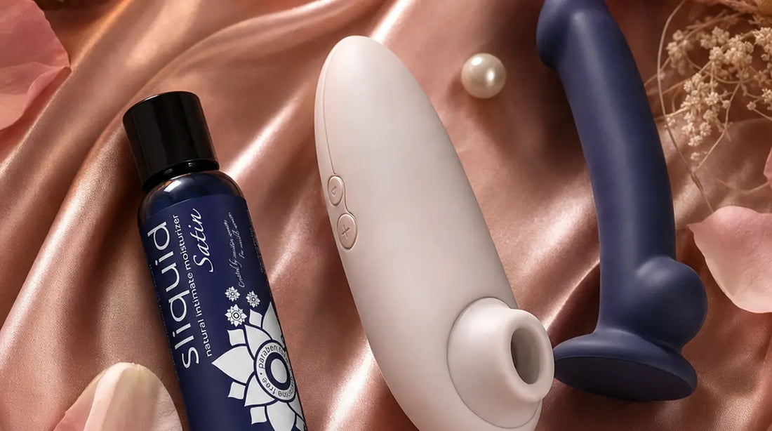 Flat-lay of a Sliquid Satin intimate moisturiser, Womanizer suction vibrator, and indigo silicone dildo arranged on blush-pink satin with rose petals, pearls, dried pampas grass, and a pink feather in warm candlelight