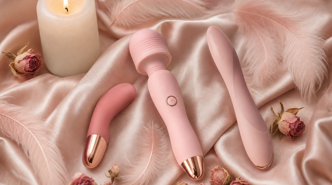 Three compact personal vibrators resting on blush-pink silk satin fabric with soft pink feathers and dried rosebuds in warm candlelight