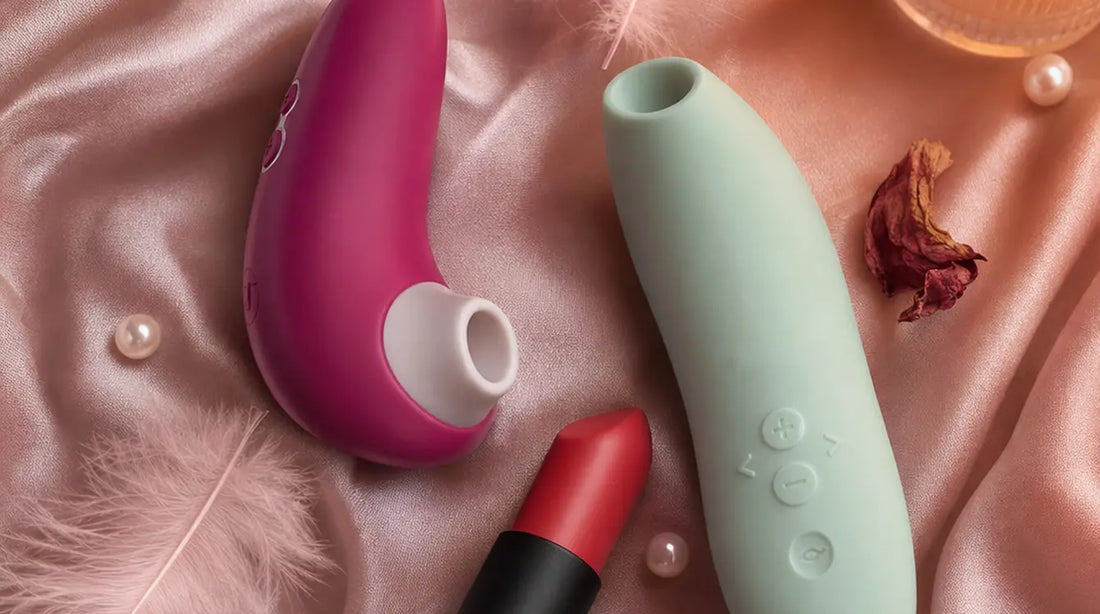 Three sleek clitoral vibrators arranged on blush-pink silk satin with pink feathers, rose petals, pearl beads, and warm candlelight
