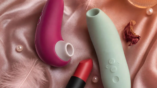 Three sleek clitoral vibrators arranged on blush-pink silk satin with pink feathers, rose petals, pearl beads, and warm candlelight
