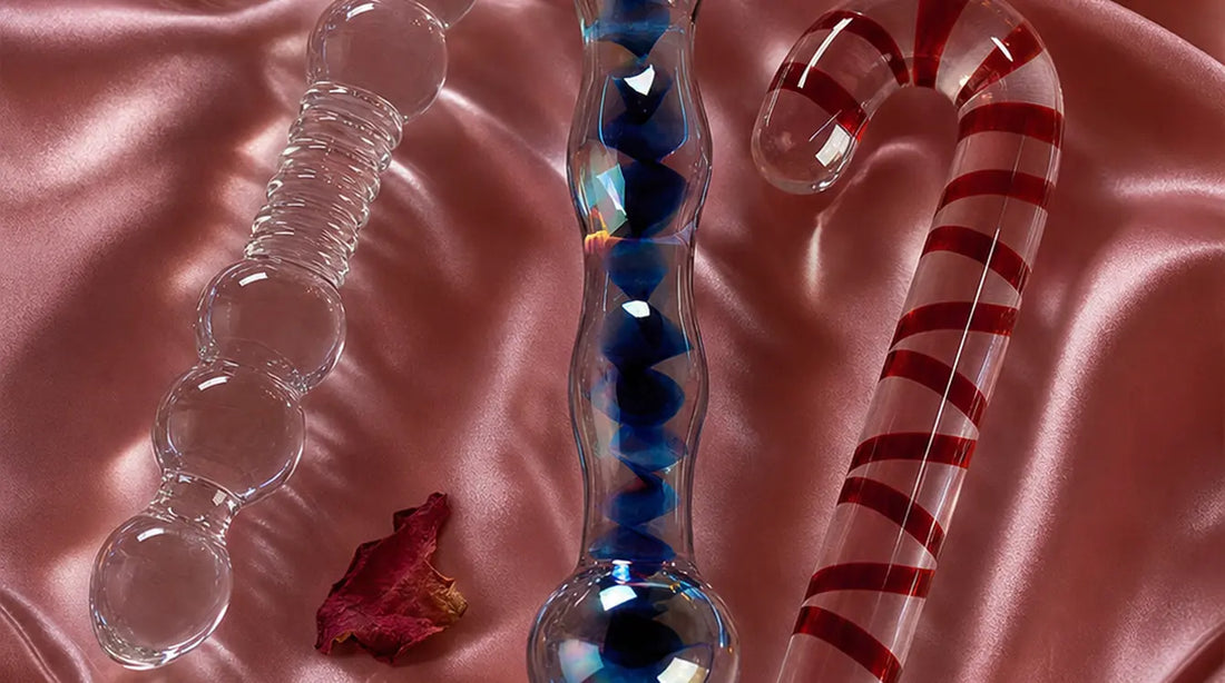 Three elegant glass dildos — clear beaded, blue-swirled, and candy-cane spiral — resting on rose-pink satin fabric with pink feathers, dried rose petals, and soft candlelight