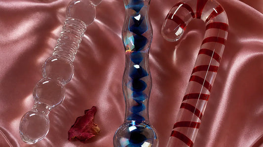 Three elegant glass dildos — clear beaded, blue-swirled, and candy-cane spiral — resting on rose-pink satin fabric with pink feathers, dried rose petals, and soft candlelight
