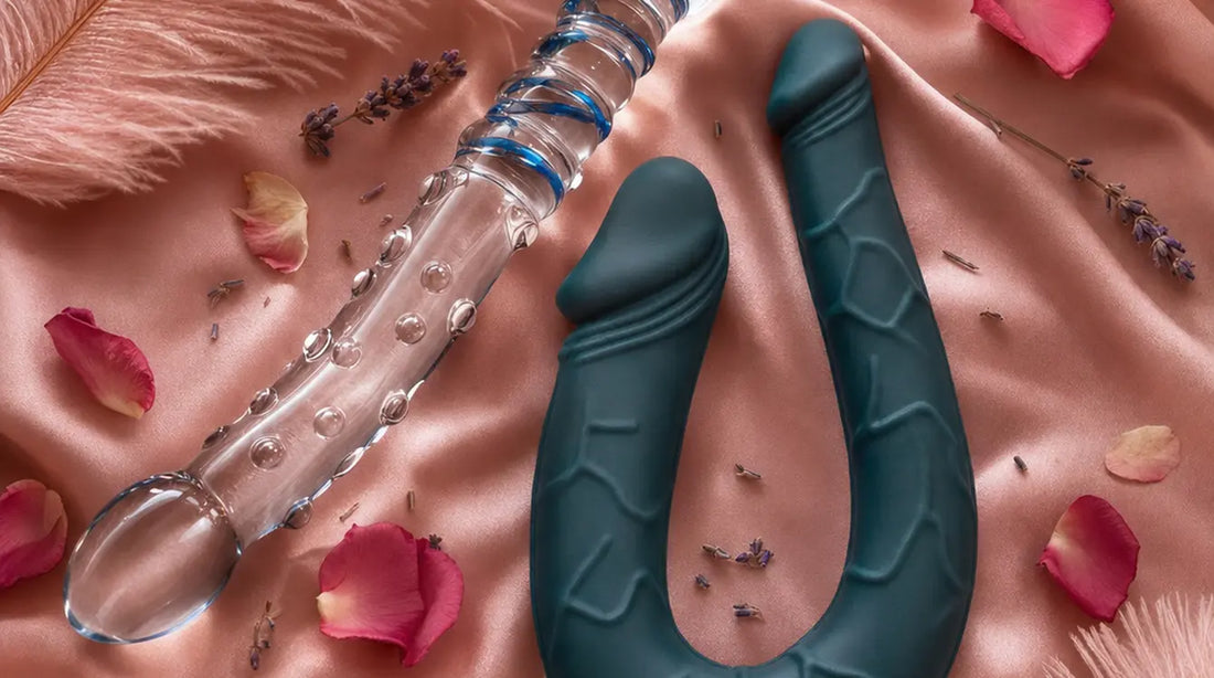 Two double-ended dildos — a clear glass spiral wand and a dark teal U-shaped silicone double dong — arranged on blush pink satin with feathers, rose petals, dried lavender, and glowing candles