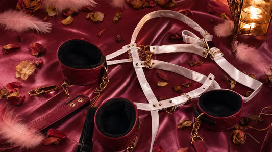 Soft padded wrist cuffs and a white satin harness with gold hardware arranged on deep rose satin fabric with pink feathers and dried rose petals, lit by warm candlelight