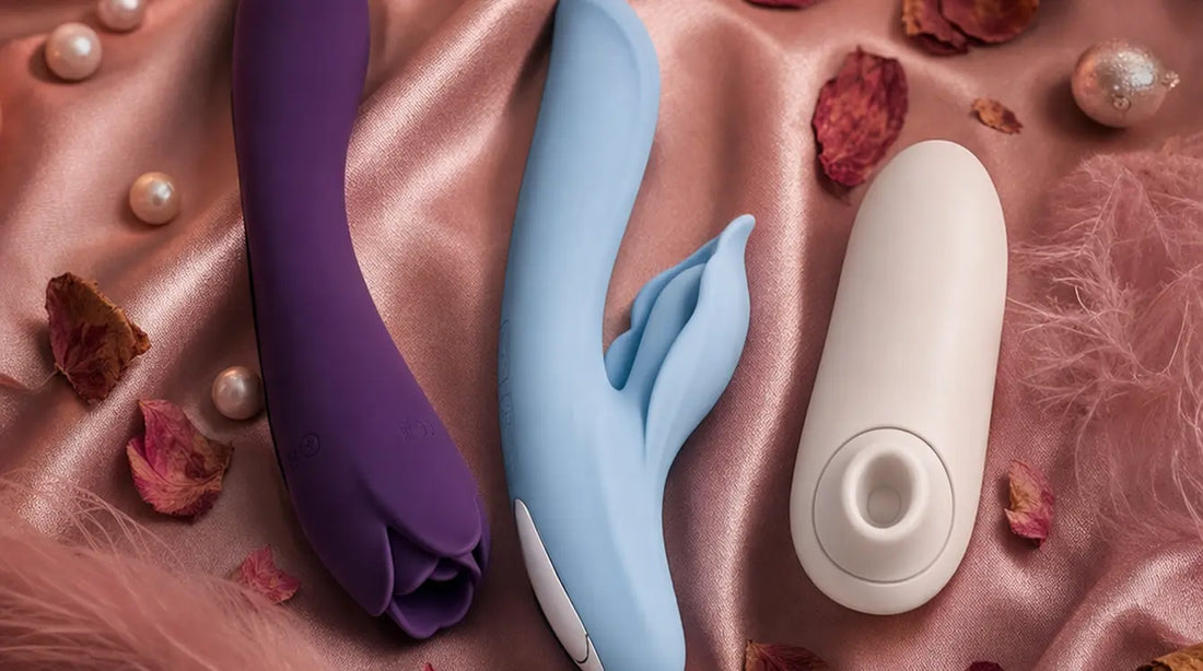 Three intimate wellness toys arranged on blush-pink silk satin with pink feathers, rose petals, pearl accents, and warm candlelight — a romantic editorial scene featuring rose-shaped and clitoral air-pulse stimulators