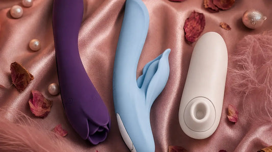 Three intimate wellness toys arranged on blush-pink silk satin with pink feathers, rose petals, pearl accents, and warm candlelight — a romantic editorial scene featuring rose-shaped and clitoral air-pulse stimulators