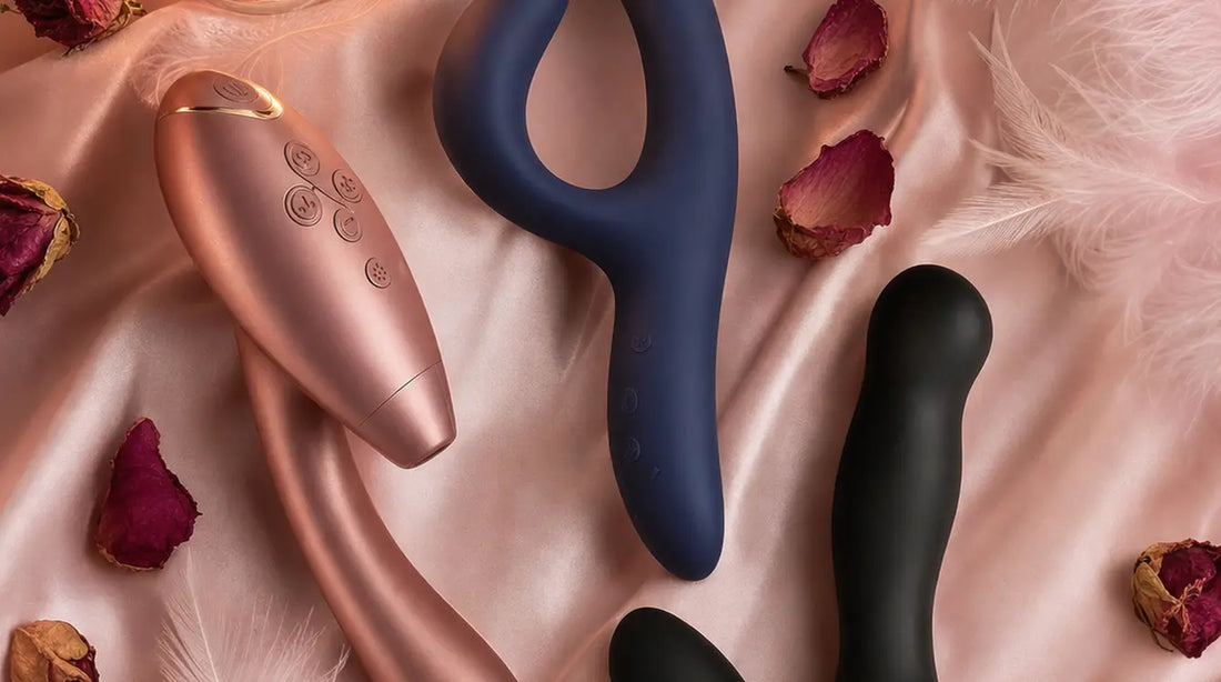 Three premium vibrators arranged on blush pink silk satin with feathers, rose petals, dried flowers, and a glowing candle in soft warm light
