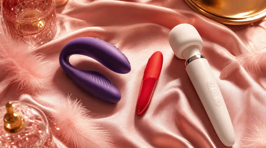We-Vibe Chorus couples vibrator, Tango X bullet, and Wand 2 on blush pink silk with feathers — brand comparison hero image