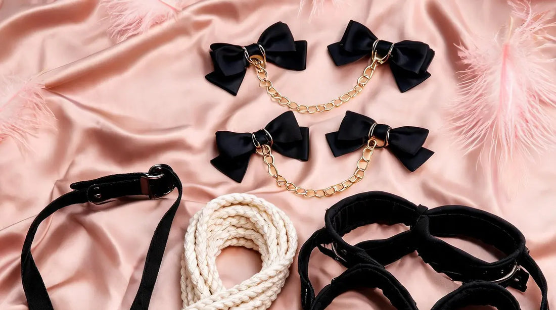 A romantic flat-lay of soft restraint products on blush-pink silk with pink ostrich feathers — black satin bow cuffs with gold chain, soft cotton rope, padded fabric cuffs, and a restraint strap with buckle