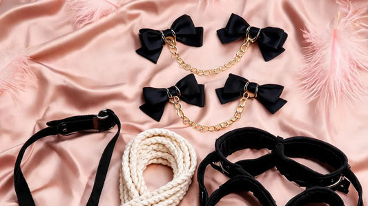 A romantic flat-lay of soft restraint products on blush-pink silk with pink ostrich feathers — black satin bow cuffs with gold chain, soft cotton rope, padded fabric cuffs, and a restraint strap with buckle