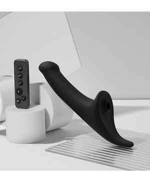 Strap-On-Me Multi Orgasm Vibrating Dildo in black shown upright with its remote control in a styled studio setting, no packaging shown